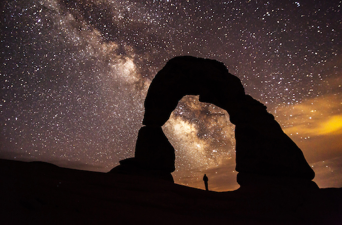 Person standing under a natural arch at Arches National Park with the Milky Way over head